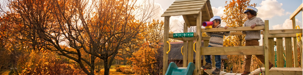 A wooden children's playground structure with a small slide and climbing platform in an outdoor park during autumn. Two children are playing on the right side, with orange trees and a soft-focus background on the left.