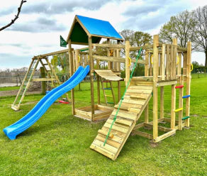 Gran parque infantil de madera con tobogán, muro de escalada y puente oscilante sobre un césped