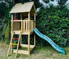 Casita de madera compacta con tobogán verde y escalera de escalada junto a un seto
